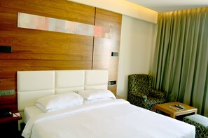 A hotel room with a large bed, a wooden headboard, a green patterned armchair, a small wooden table, and a green curtain. - R K Residency (Tirupur)