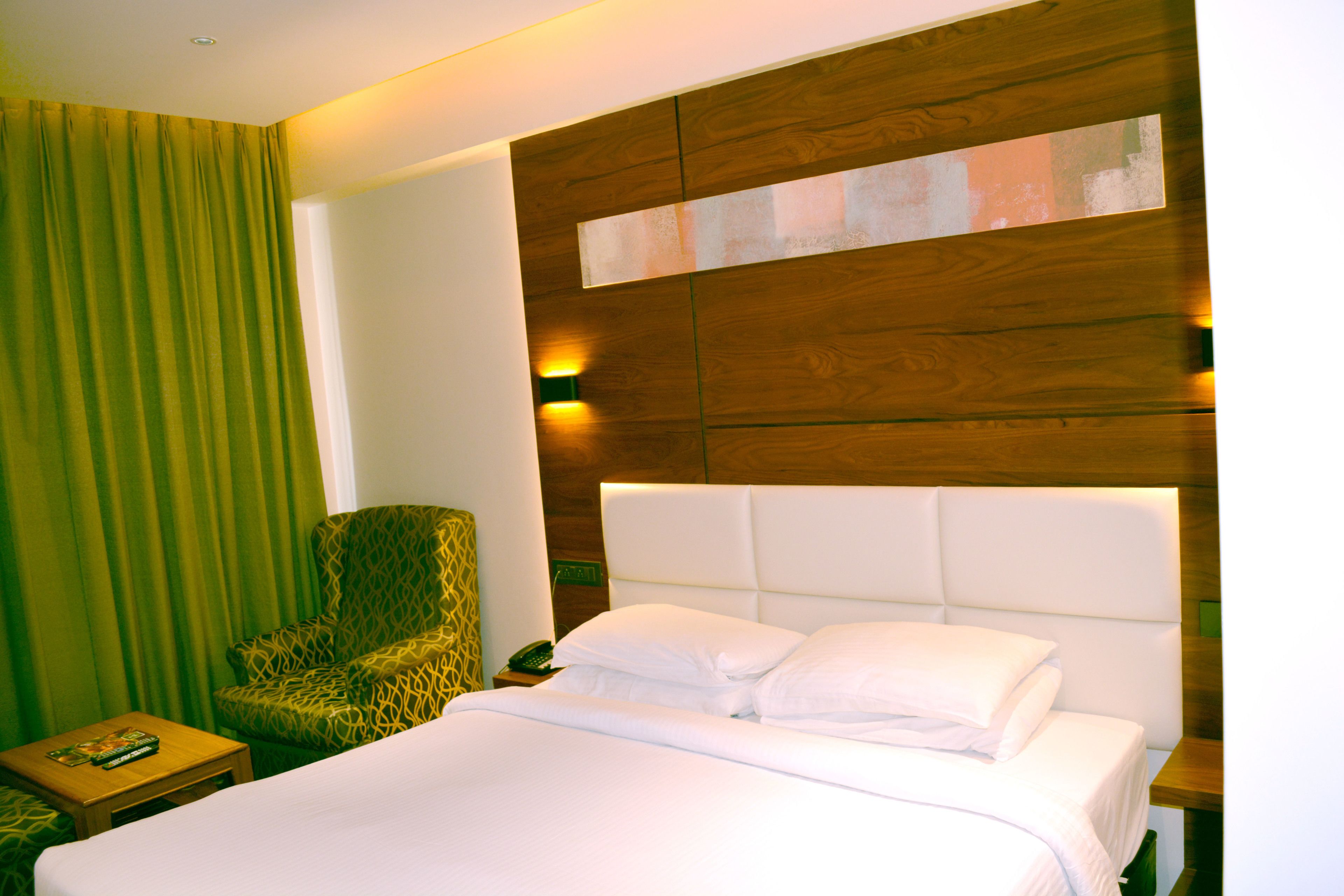 A hotel room with a large bed, a patterned armchair, a bedside table with a lamp, and a wooden headboard with a light panel.