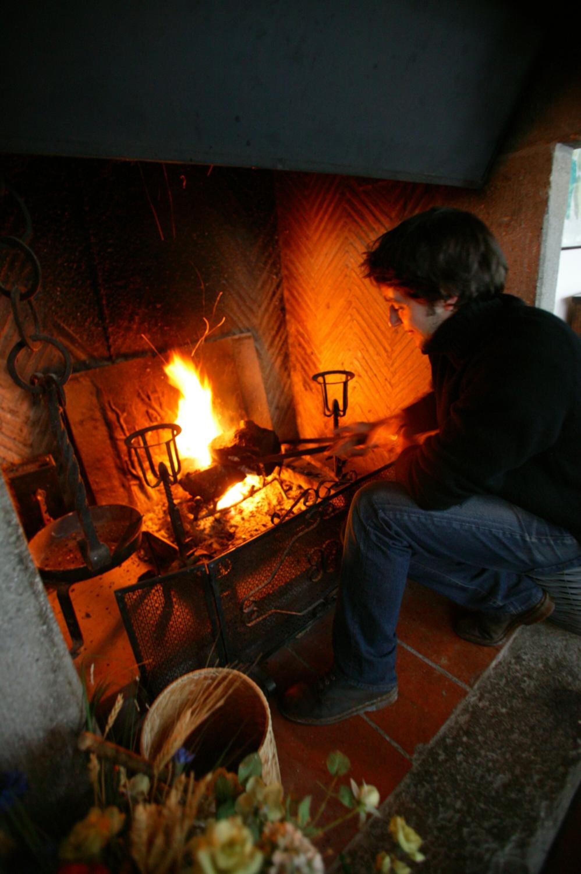 Une personne est assise près d’une cheminée où brûle un feu, entourée de divers objets, comme une marmite et un panier.