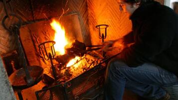 Une personne est assise près d’une cheminée où brûle un feu, entourée de divers objets, comme une marmite et un panier.