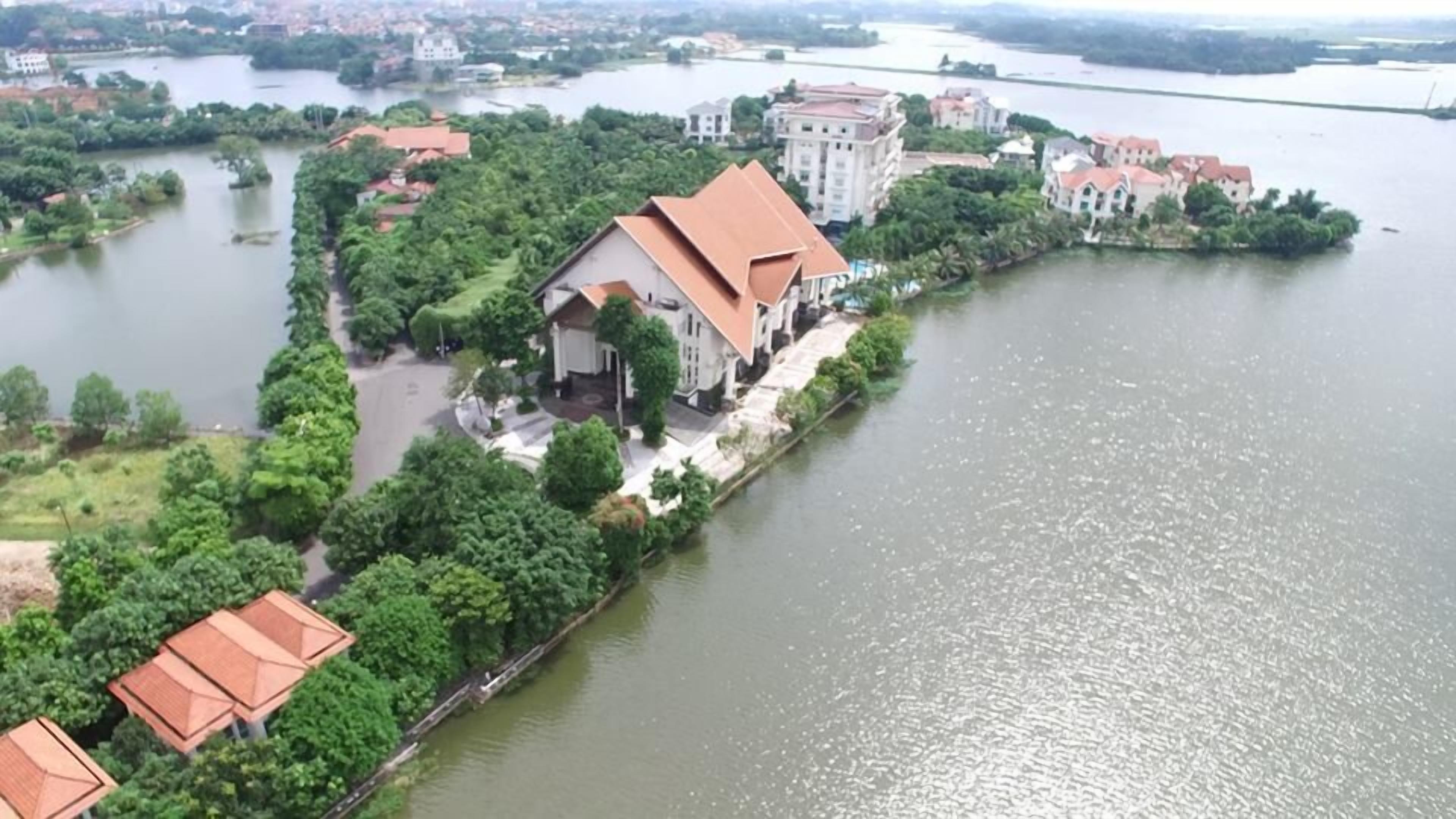 An aerial view of a riverside property with a prominent building featuring a red roof, surrounded by lush greenery and adjacent to a calm river.
