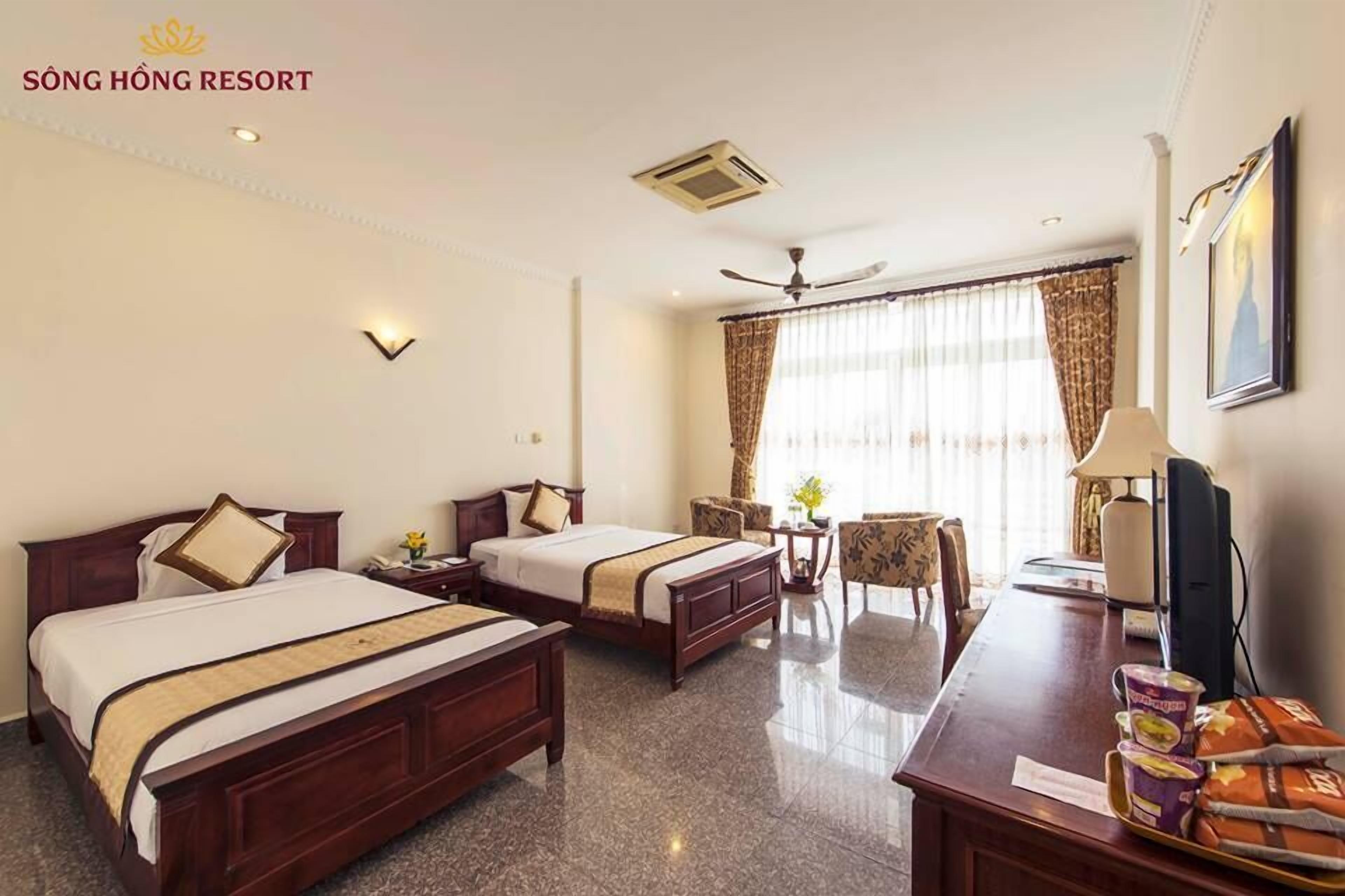 A hotel room with two beds, a desk with a computer, a chair, a table with a lamp, and a window with curtains.