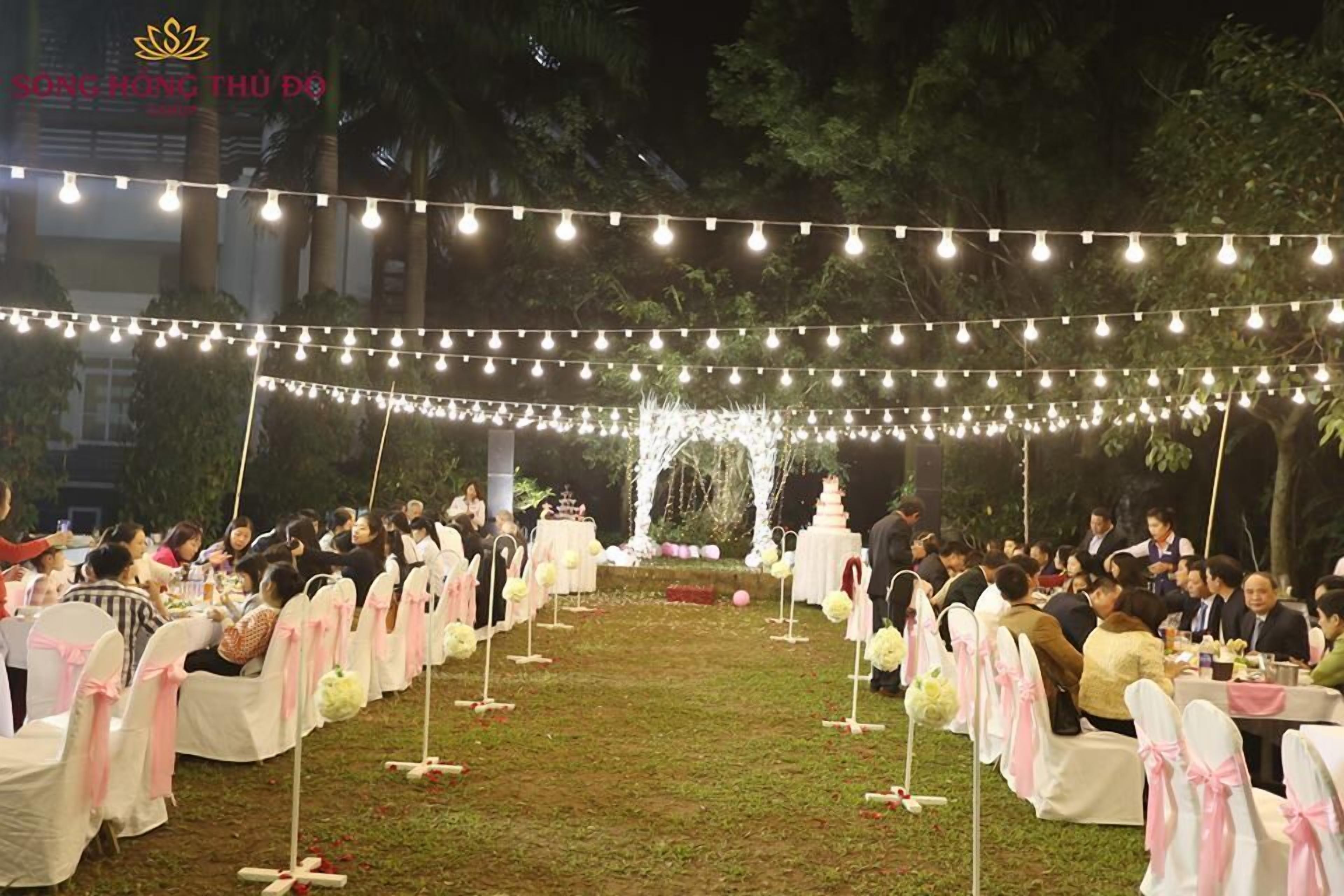 An outdoor event with string lights overhead, tables with white tablecloths, and chairs with pink sashes.