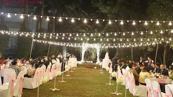 An outdoor event with string lights overhead, tables with white tablecloths, and chairs with pink sashes.