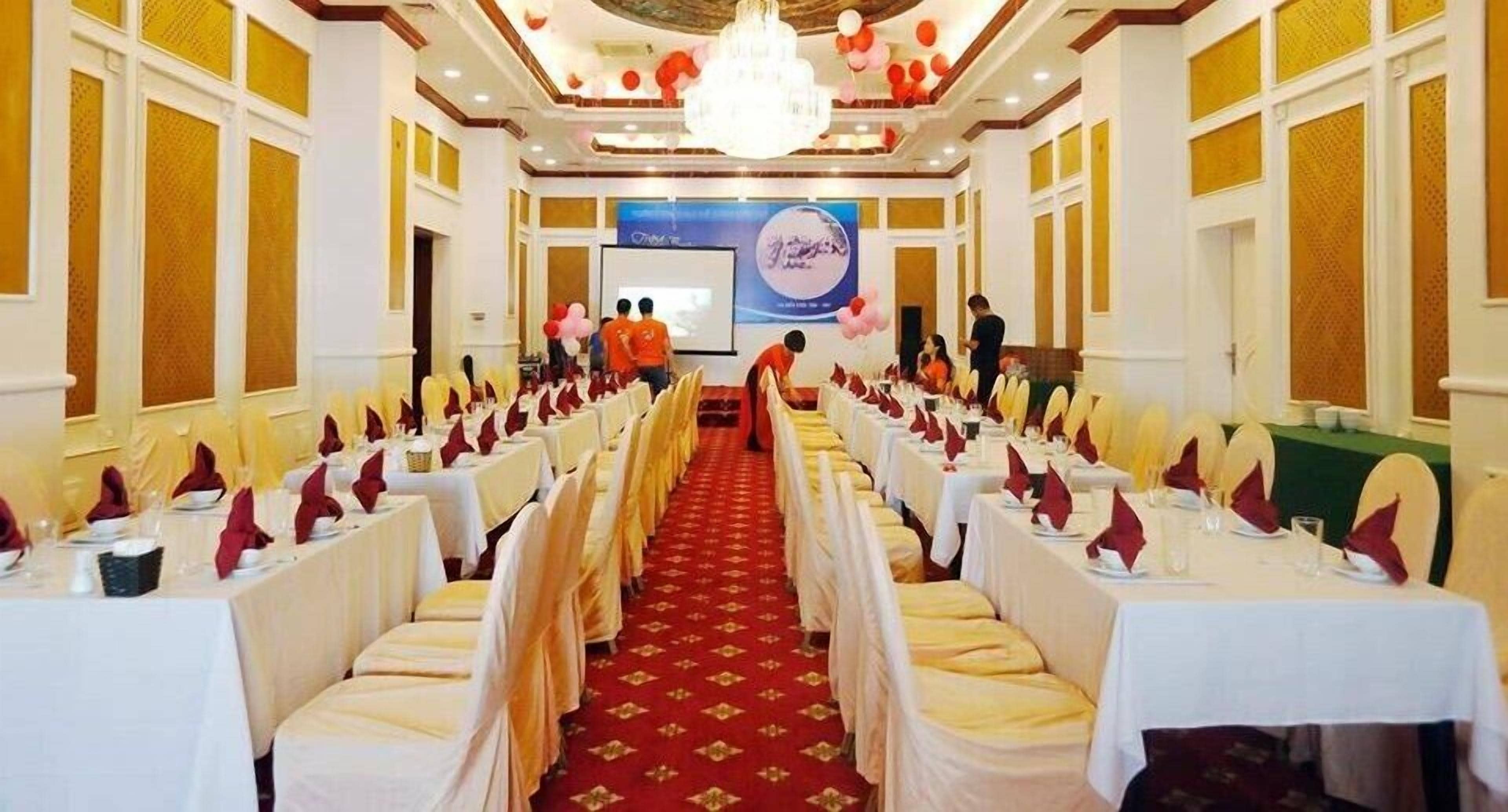 A banquet hall with rows of tables set for a formal event, a stage with a presentation screen, and a chandelier.