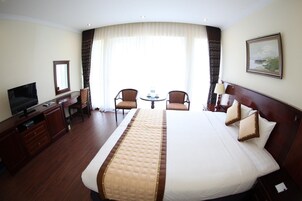 Quarto de hotel com uma cama grande, uma escrivaninha com televisão, duas cadeiras, uma mesa pequena e um quadro na parede.