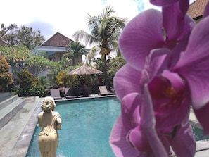 A pool area with a statue, a palm tree, and a building in the background. - Kartika Bungalows (Ubud)