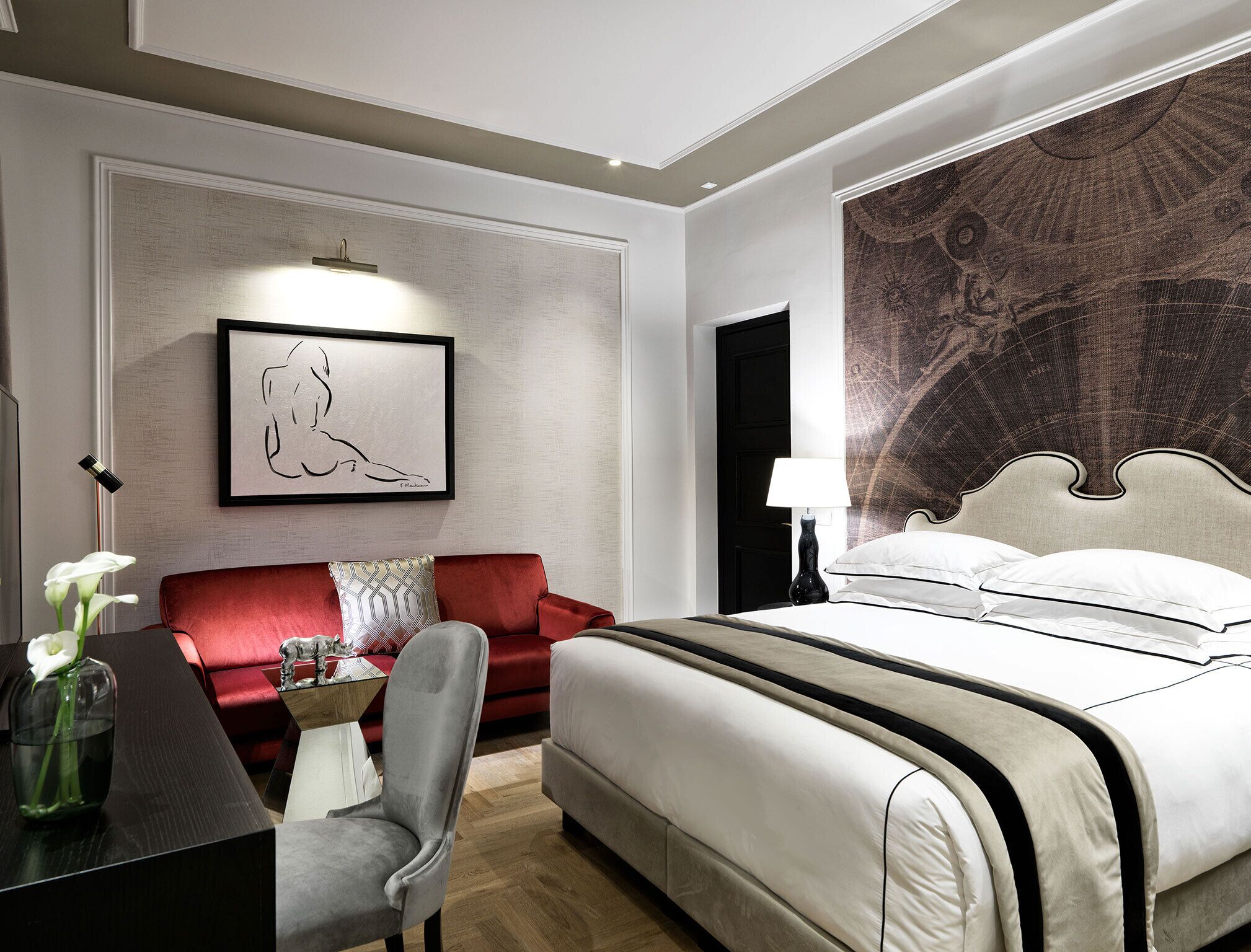 A hotel room with a bed, a red sofa, a desk with a chair, a framed artwork on the wall, and a large tapestry.