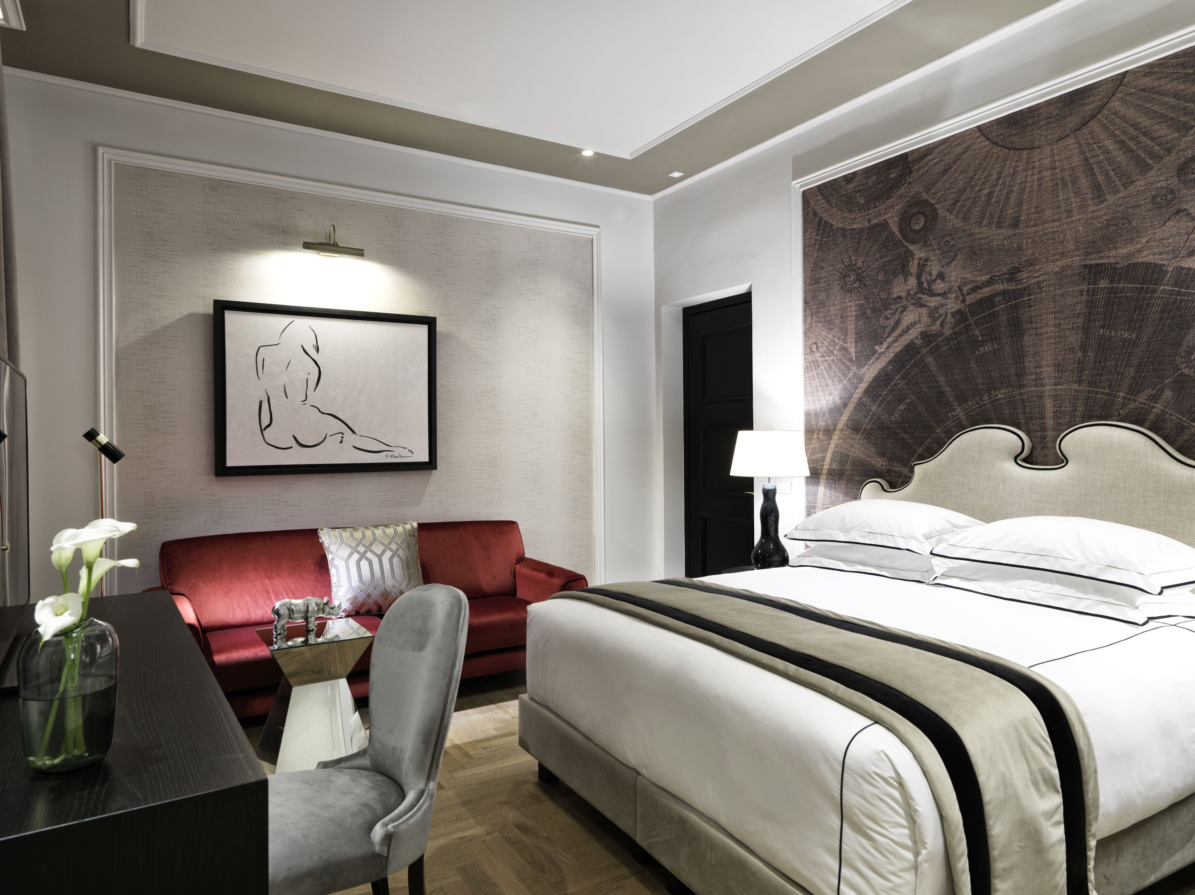 Een moderne hotelkamer met een groot bed, een rode bank, een kleine tafel met een vaas, en een wand met een groot wandtapijt.