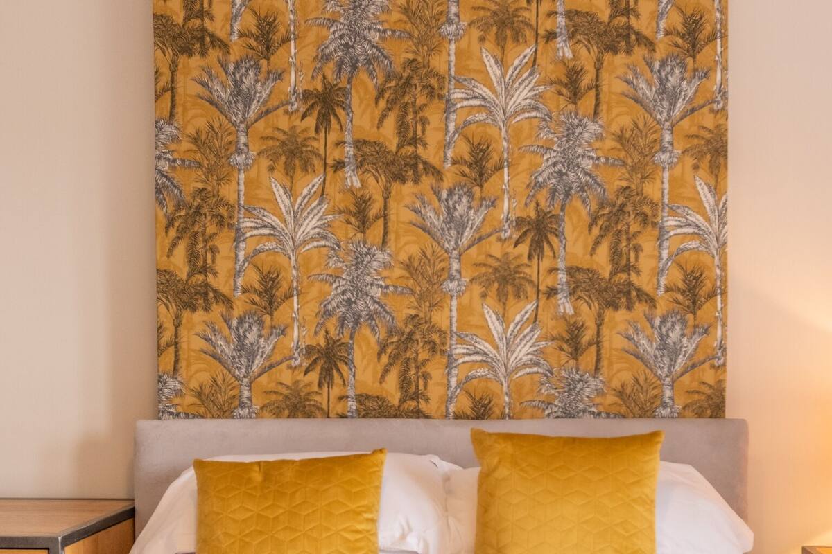 Una cama bien tendida con dos almohadas amarillas, una revista sobre la cama y un tapiz grande con estampado de hojas tropicales en la pared.