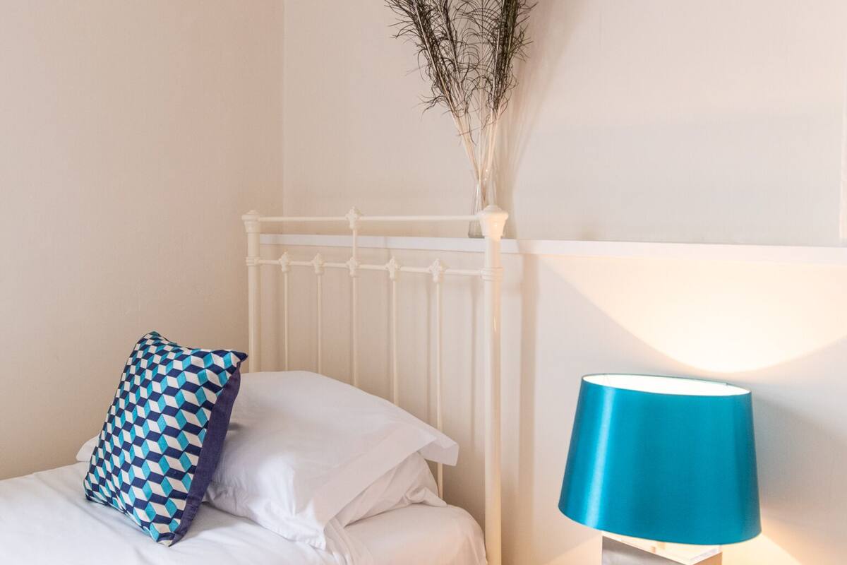 Una cama bien hecha con un cojín estampado, una mesita de noche con una lámpara y un jarrón con plantas secas en la pared.