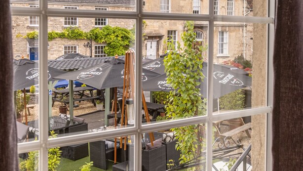 A view through a window of a courtyard with outdoor seating, potted plants, and stone buildings in the background. - Rose And Crown (Bath)