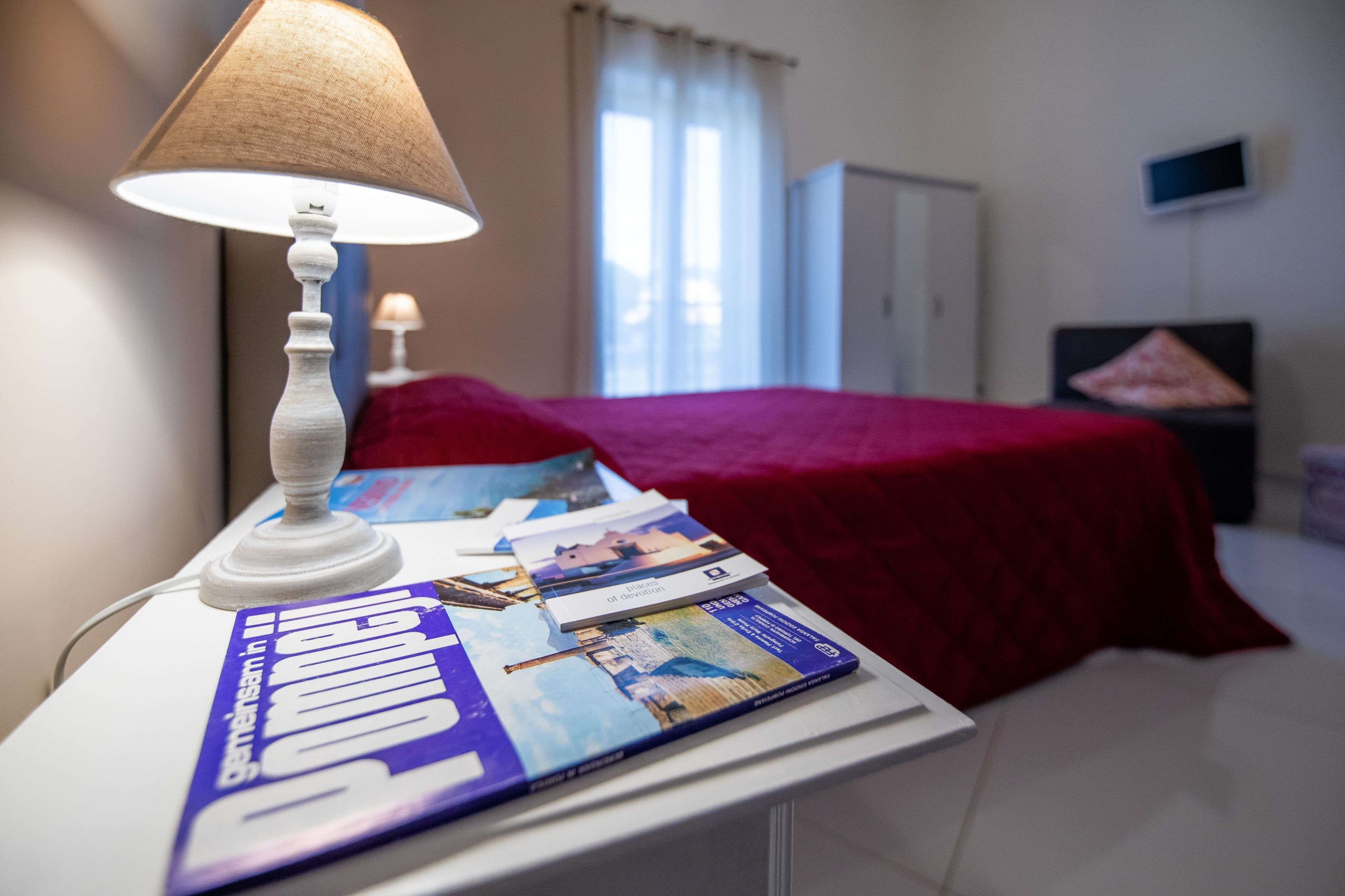A hotel room with a bed covered in a red blanket, a white lamp on a bedside table, and a magazine on the table.