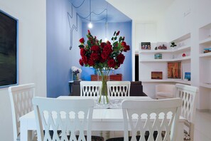Un coin repas avec une table et des chaises blanches, un vase de roses rouges et un mur d’accent bleu.