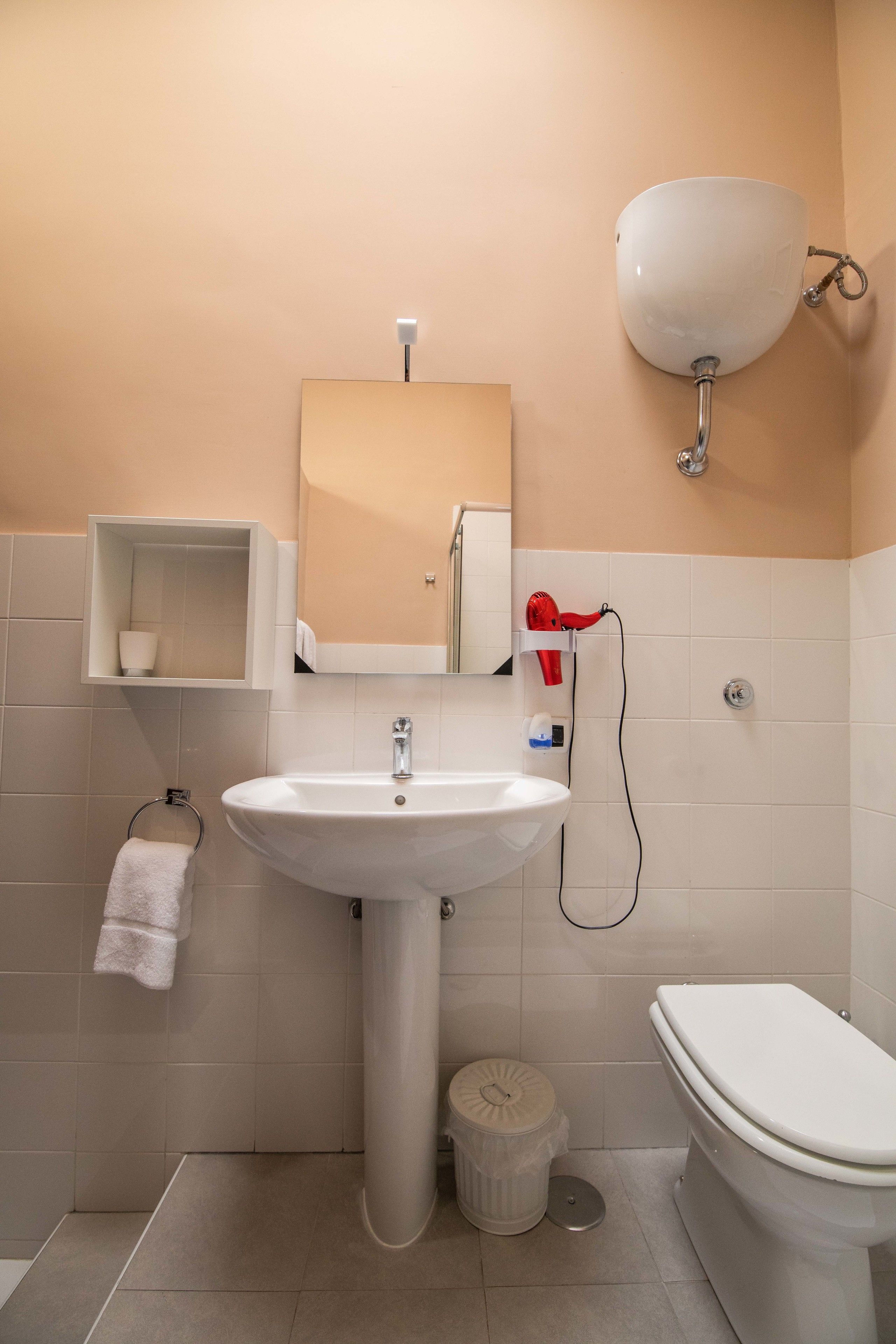 A bathroom with a pedestal sink, a toilet, a mirror, a towel rack, and a red hairdryer.