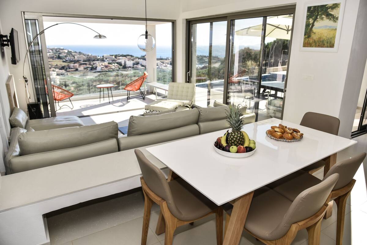 A modern living room with a large window, a sofa, a dining table with fruit, and a view of the sea.