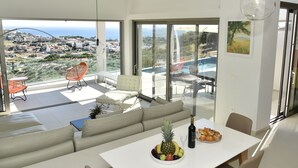 A modern living room with a sofa, dining table, and a view of a pool and cityscape. - Sea View Villas (Malevizi)
