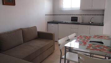 Un espacio habitable compacto con sofá, mesa de comedor con sillas, kitchenette con microondas y ventana con persianas.