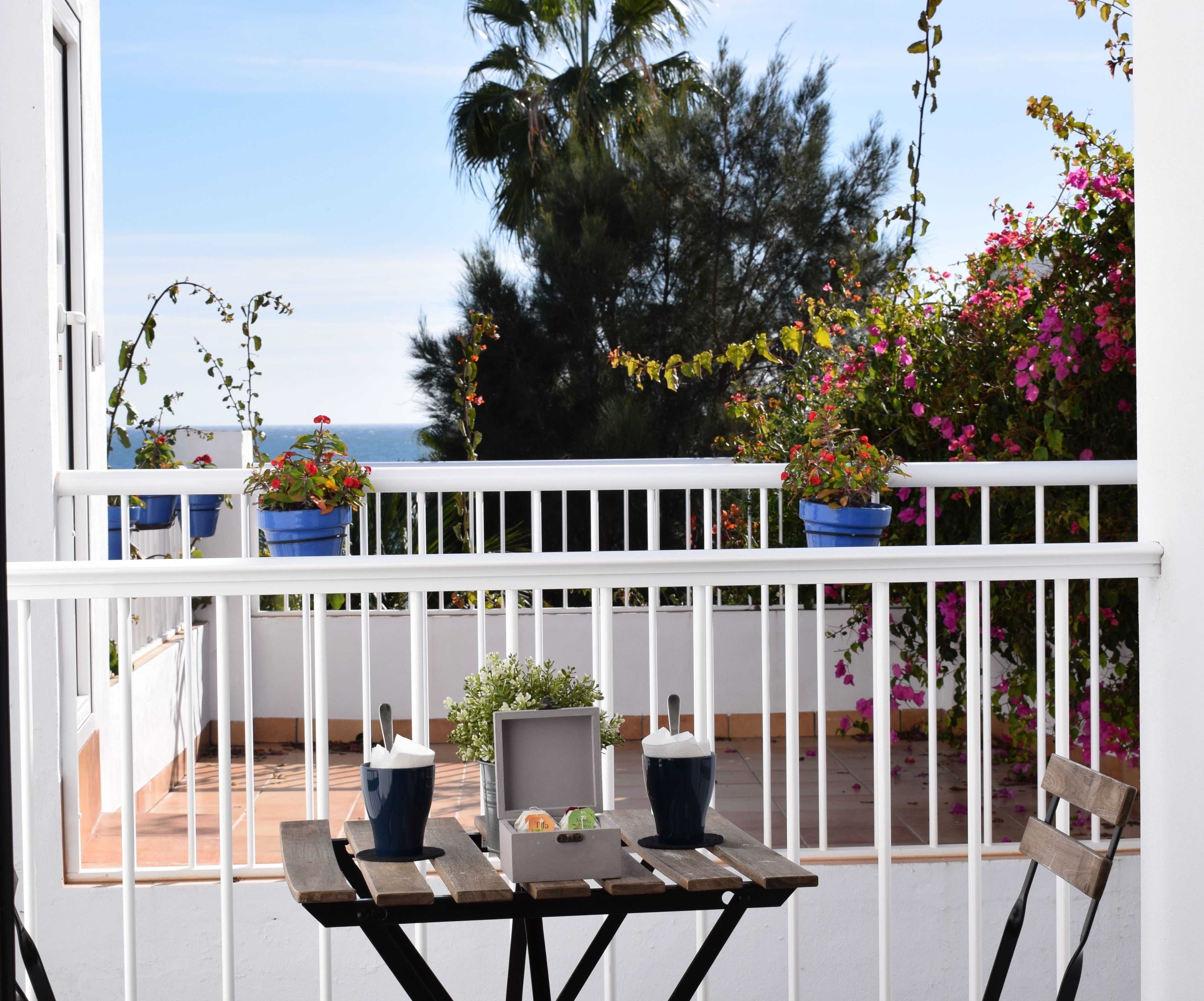 Een balkon met een witte balustrade, plantenpotten, een tafel met stoelen en uitzicht op de oceaan.