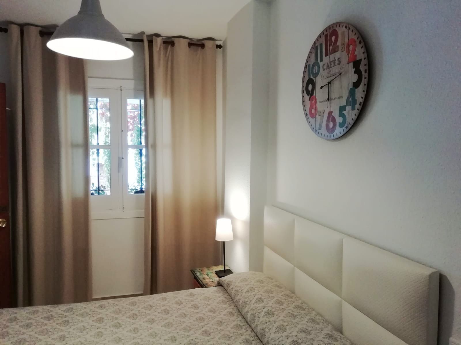 Een slaapkamer met een bed, een wandklok, een raam met gordijnen en een hanglamp.