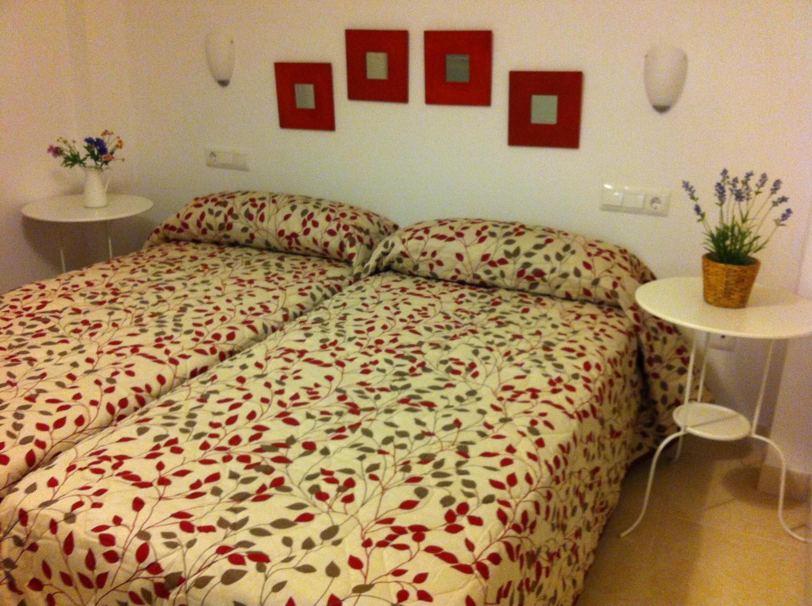 Una cama doble con colcha estampada de flores, dos mesitas de noche con lámparas y una pequeña planta en maceta sobre una de ellas.