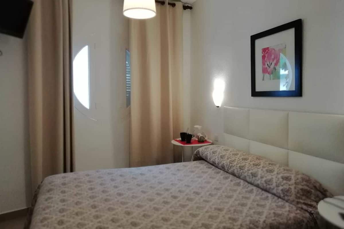A hotel room with a bed, a bedside table with a lamp, a framed picture on the wall, and a window with curtains.