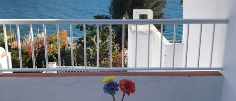 A balcony with a clear view of the ocean, a white railing, and a vase with flowers on a table.