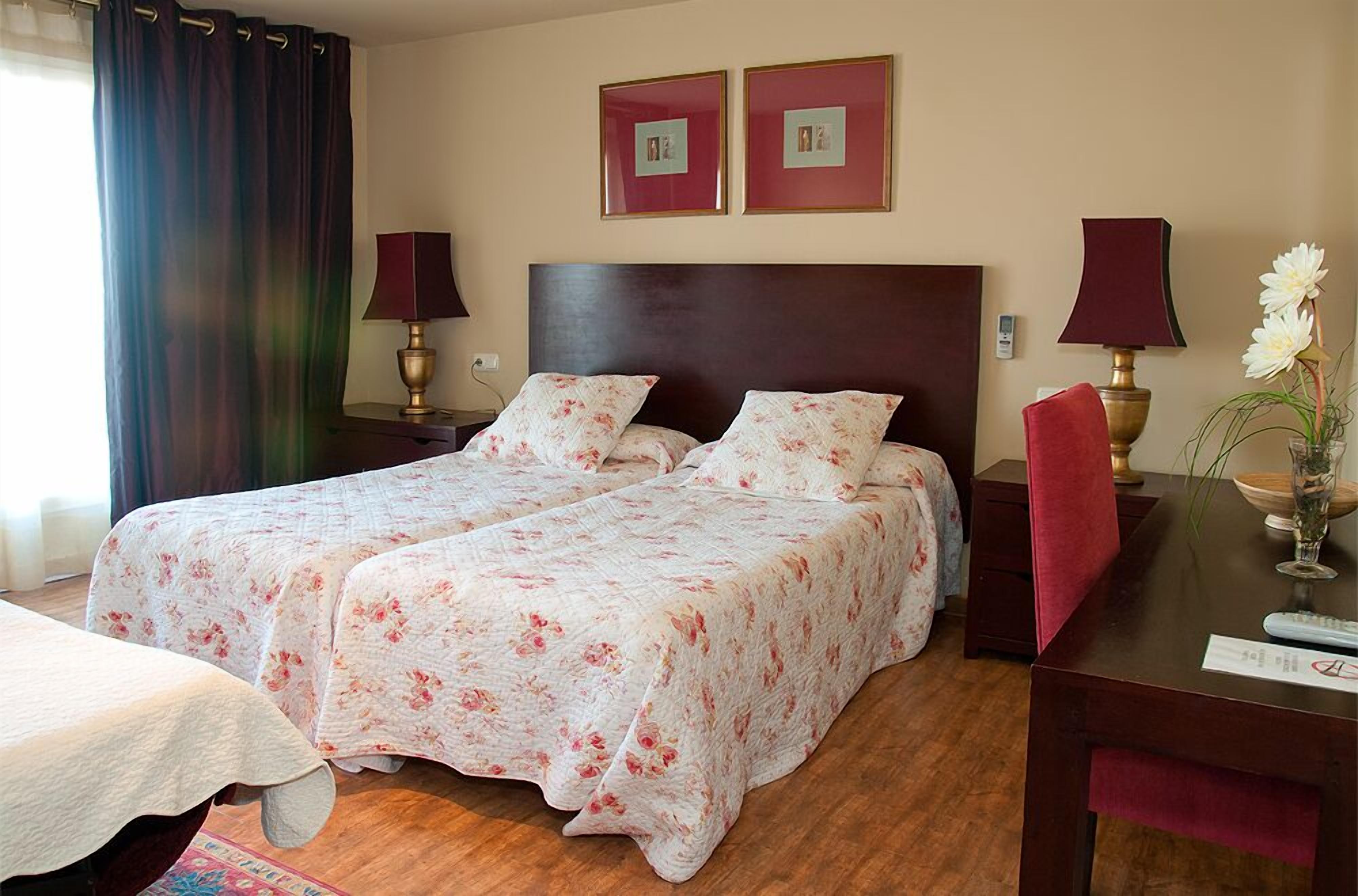 A hotel room with two beds, a wooden headboard, two bedside lamps, a desk with a vase of flowers, and two framed pictures on the wall.