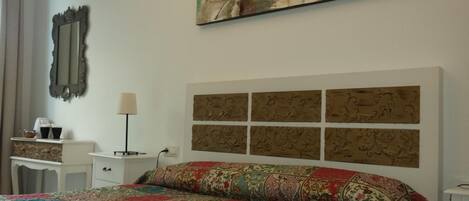 A bedroom with a large bed, a nightstand, a lamp, a mirror, and a wall-mounted artwork.