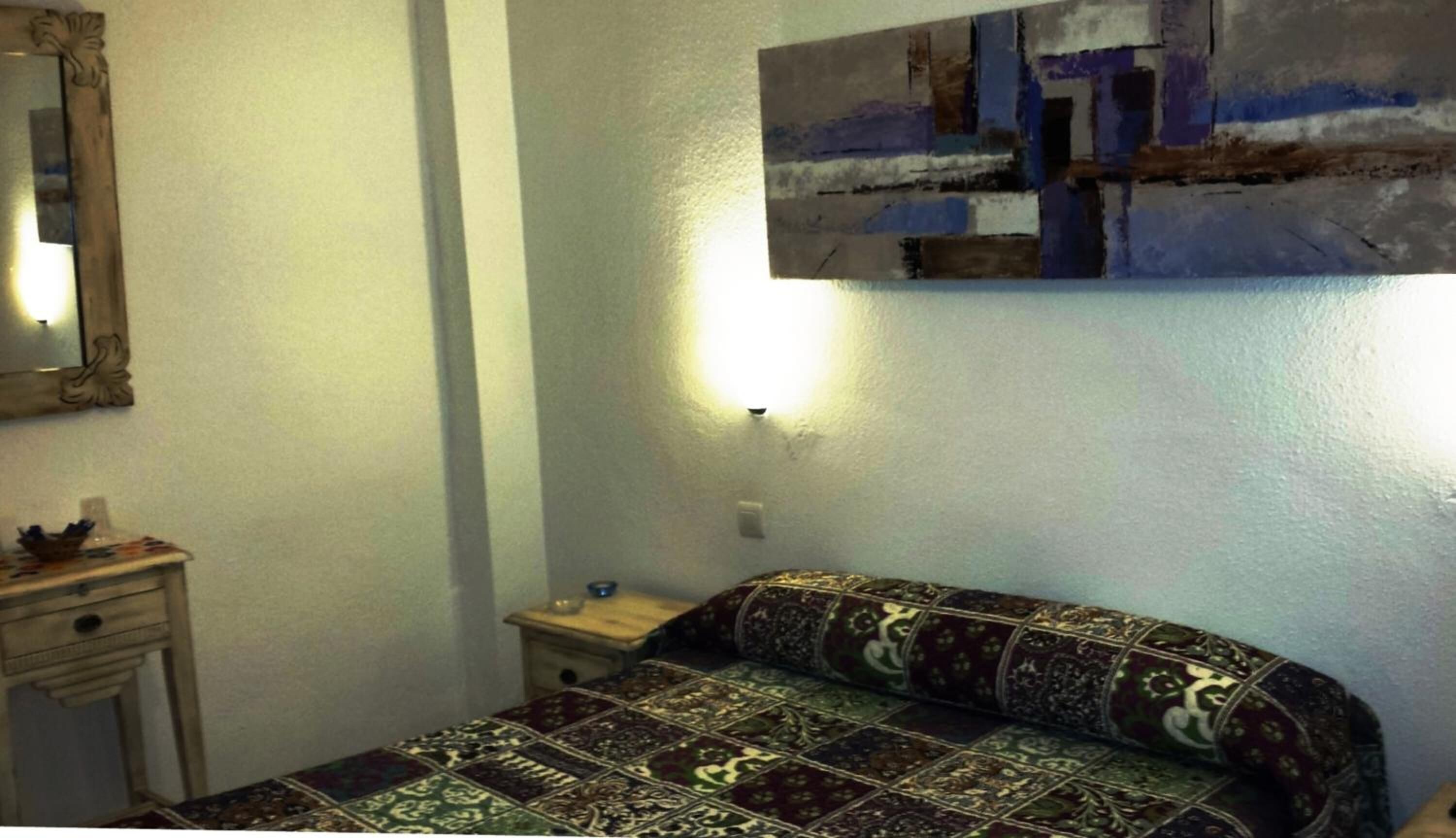 A small, simple room with a bed, a nightstand, a mirror, and a wall-mounted light.