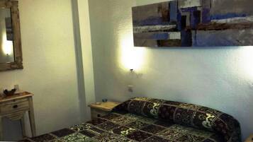 A small, simple room with a bed, a nightstand, a mirror, and a wall-mounted light.