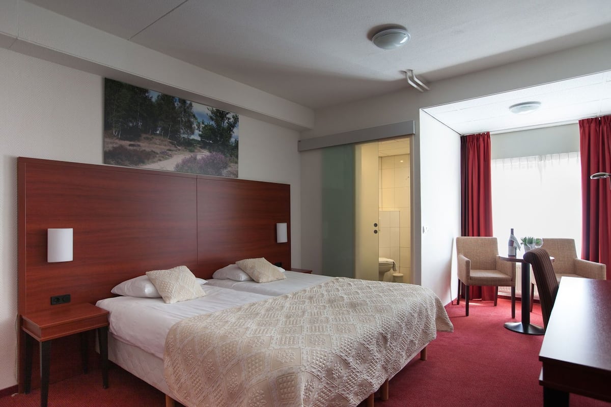 Habitación de hotel con una cama grande, un escritorio, dos sillas, un cuadro en la pared y un baño visible a través de una puerta abierta.