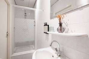 Un baño moderno con ducha envuelta en vidrio, un lavamanos blanco con grifo cromado y un florero con flores secas sobre un estante blanco.
