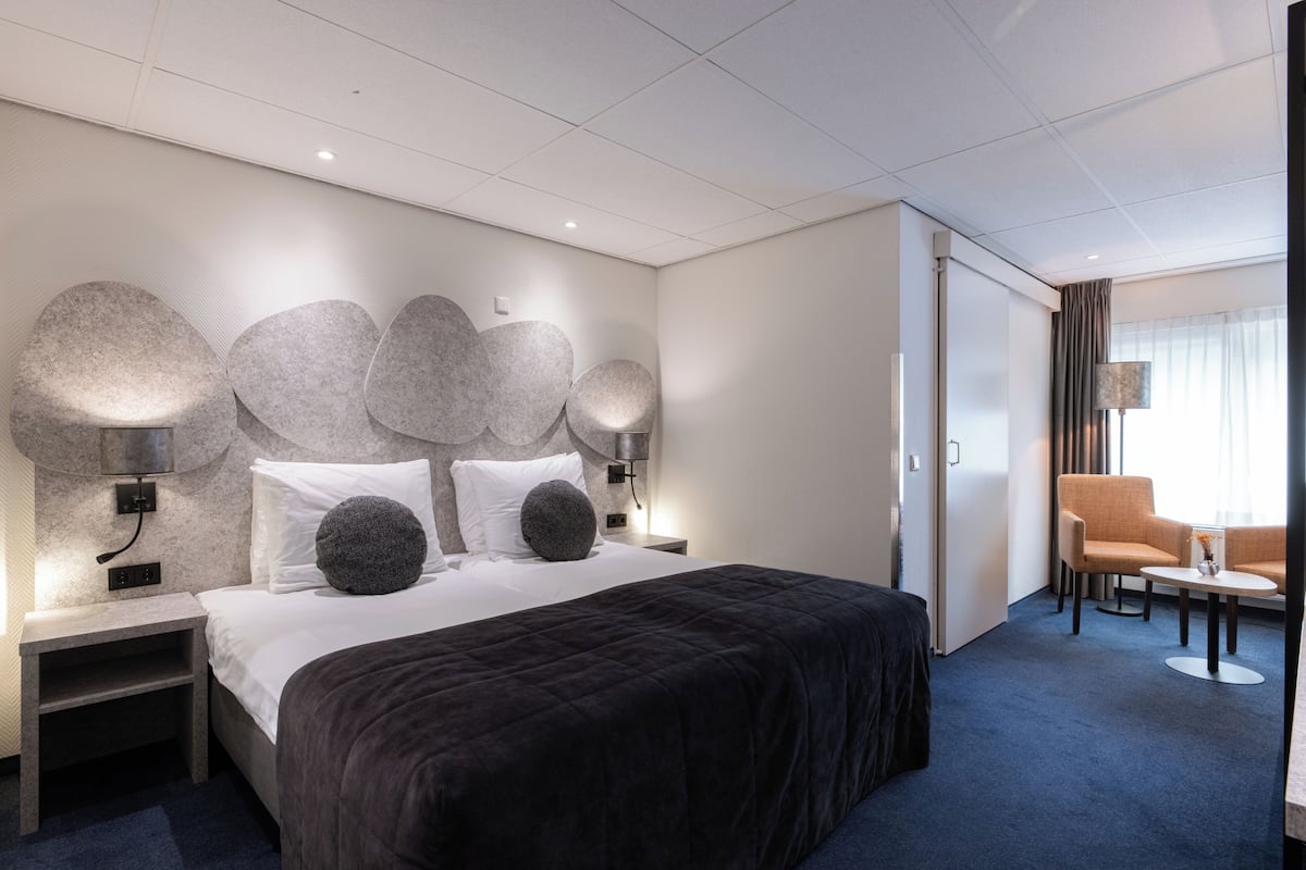 Una habitación de hotel moderna con una cama grande, dos mesitas de noche, un escritorio con silla y una zona de estar con sofá y sillones.