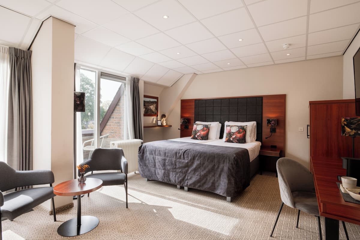 Una habitación de hotel moderna con una cama grande, un escritorio y una silla. Cuenta con una ventana con cortinas, un radiador y una pequeña mesa con un florero.