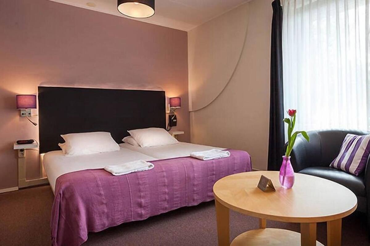 Una habitación de hotel con una cama, una silla, una mesita y un florero con una flor.