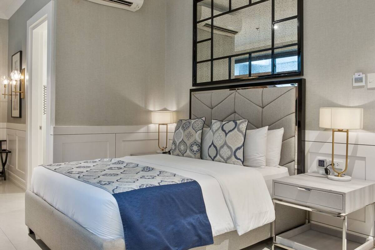 Un dormitorio moderno con una cama grande, dos mesitas de noche con lámparas, un espejo sobre la cama y vistas al exterior a través de la ventana.