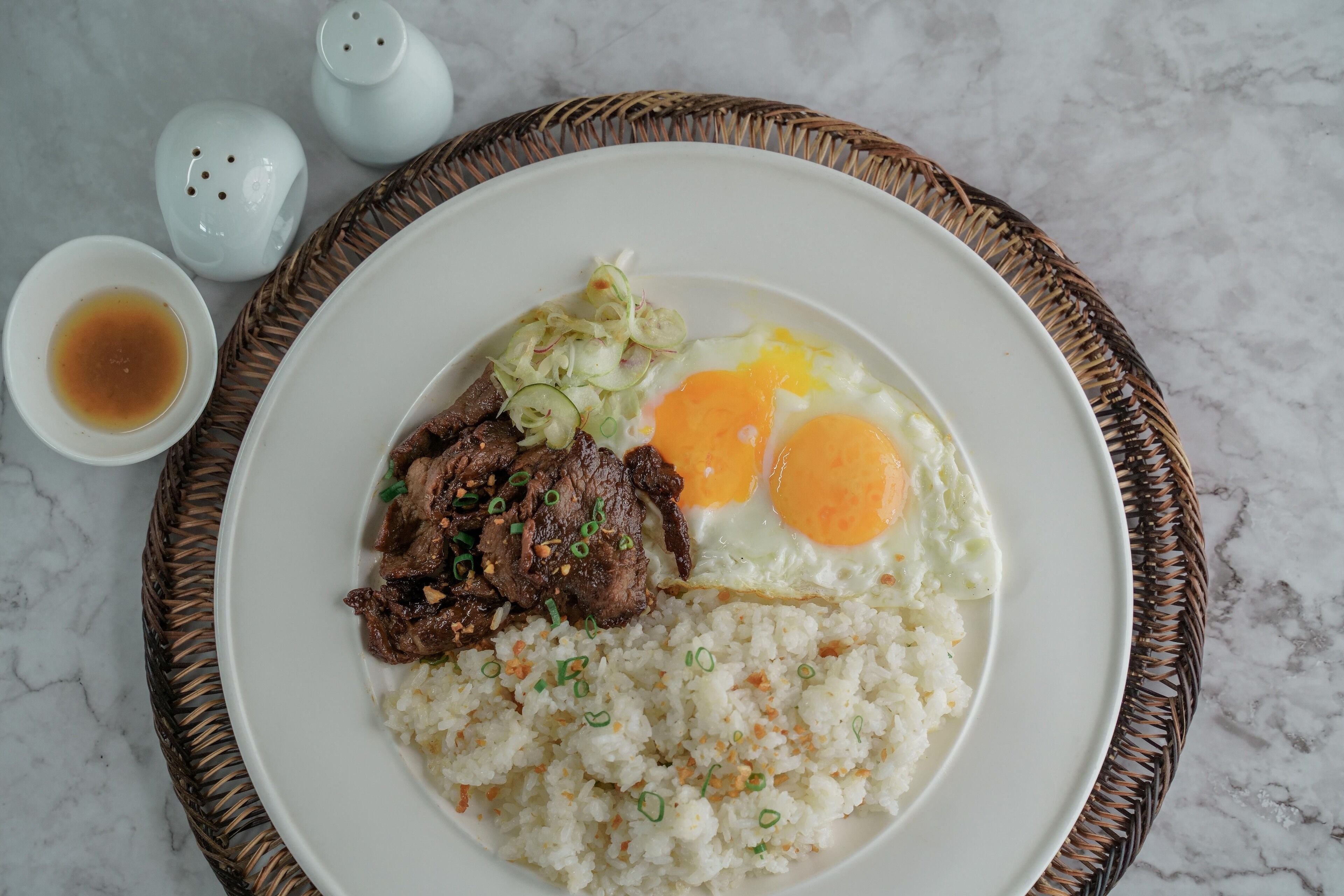 A plate of food with rice, meat, and eggs on a white plate, accompanied by a small bowl of sauce and salt and pepper shakers.