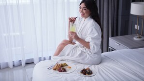 A person sitting on a bed in a hotel room with a breakfast tray and a drink. - Stonehill Suites (Bacolod)
