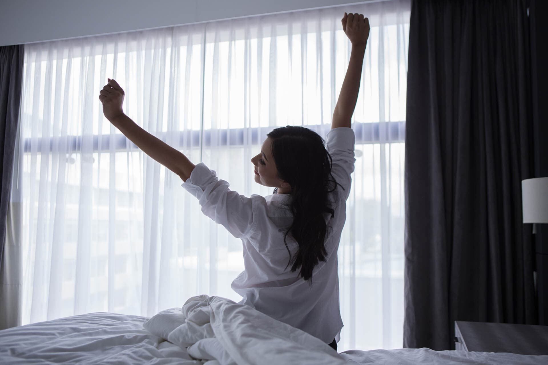 A person sitting on a bed with arms raised, in a room with a large window and curtains.