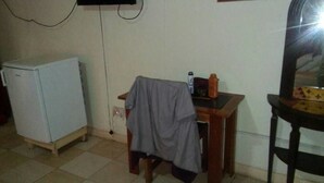 A small room with a white refrigerator, a wooden table with a jacket draped over it, and a mirror on a wooden stand. - New Mexico Hotel - Takoradi (Sekondi-Takoradi)