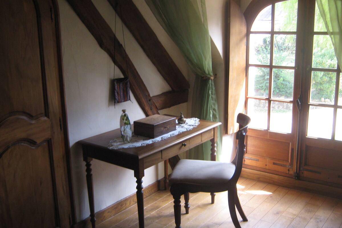 Une pièce avec un bureau en bois, une chaise, une fenêtre avec des rideaux et une vue sur la verdure.