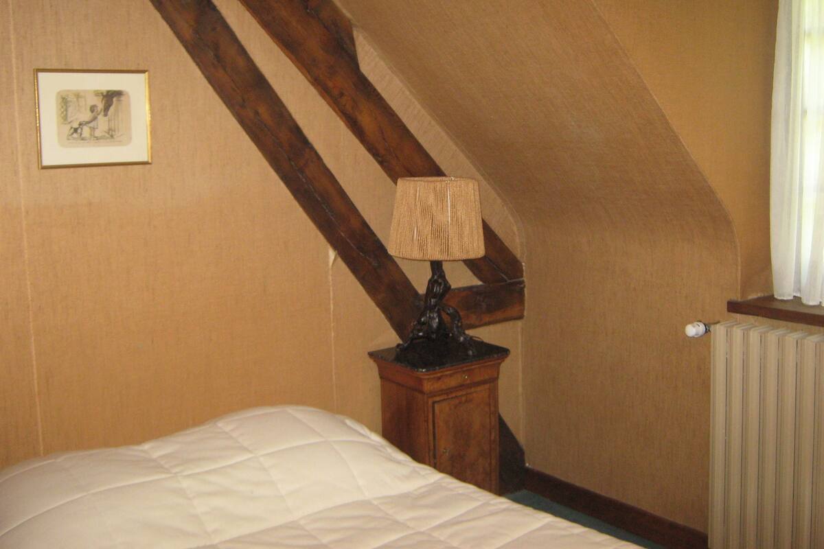 Une chambre à coucher avec un lit, une table de chevet en bois surmontée d’une lampe, un radiateur et un tableau encadré accroché au mur.