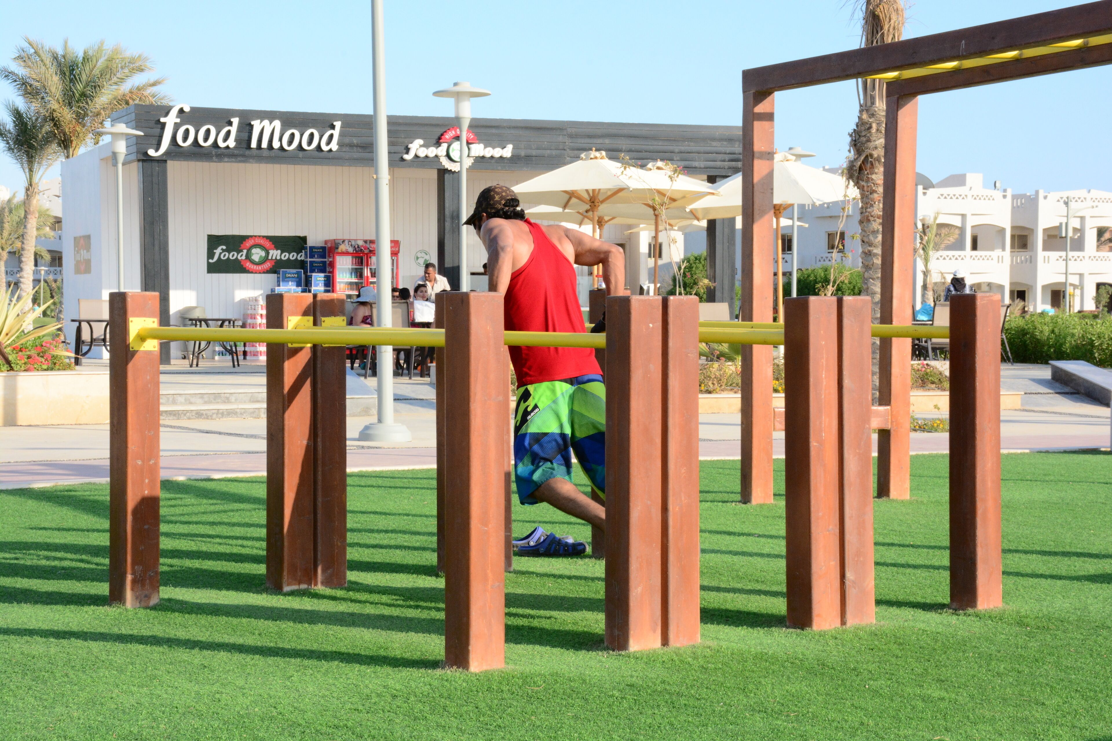 A person exercising on outdoor gym equipment in a public park area with a food court in the background.