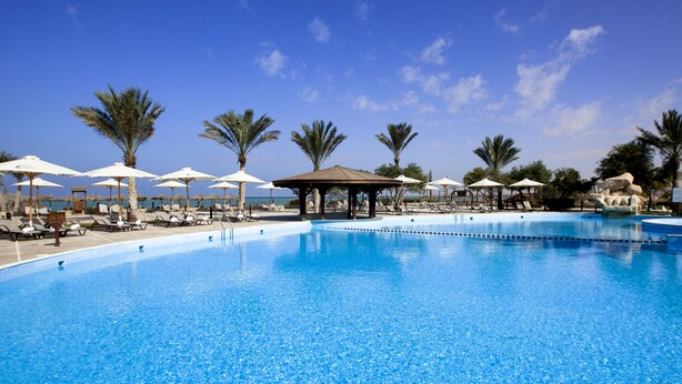 A large outdoor swimming pool with multiple lanes, surrounded by sun loungers and umbrellas, palm trees, and a clear blue sky. - Mousa Coast (Ras Sudr)
