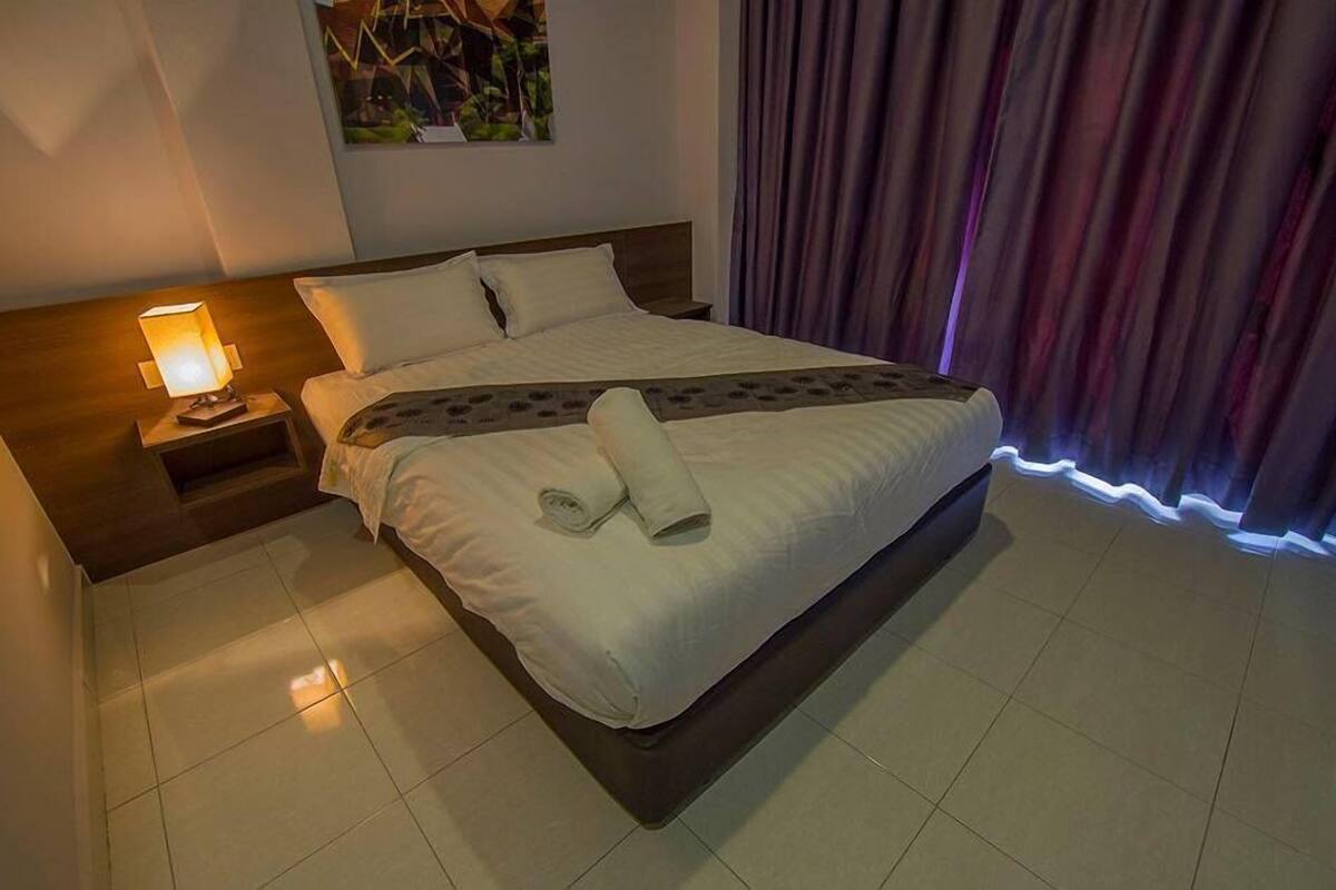 A neatly made bed with a rolled towel, a nightstand with a lamp, and a painting on the wall.