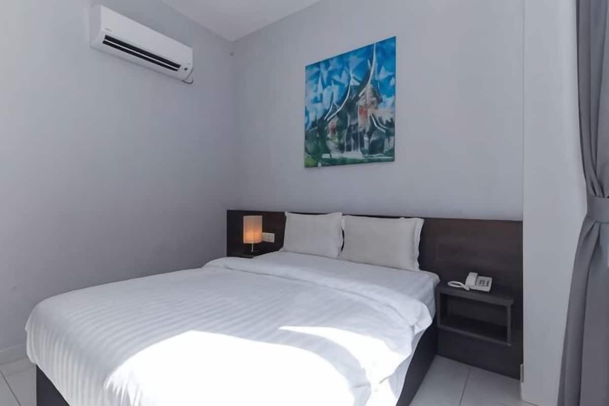 A bedroom with a bed, an air conditioner, a nightstand with a lamp, and a painting on the wall.