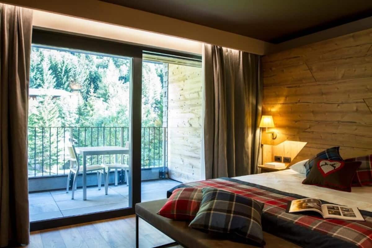 A bedroom with a large bed, a nightstand with a lamp, and a view of a balcony with a table and chairs.