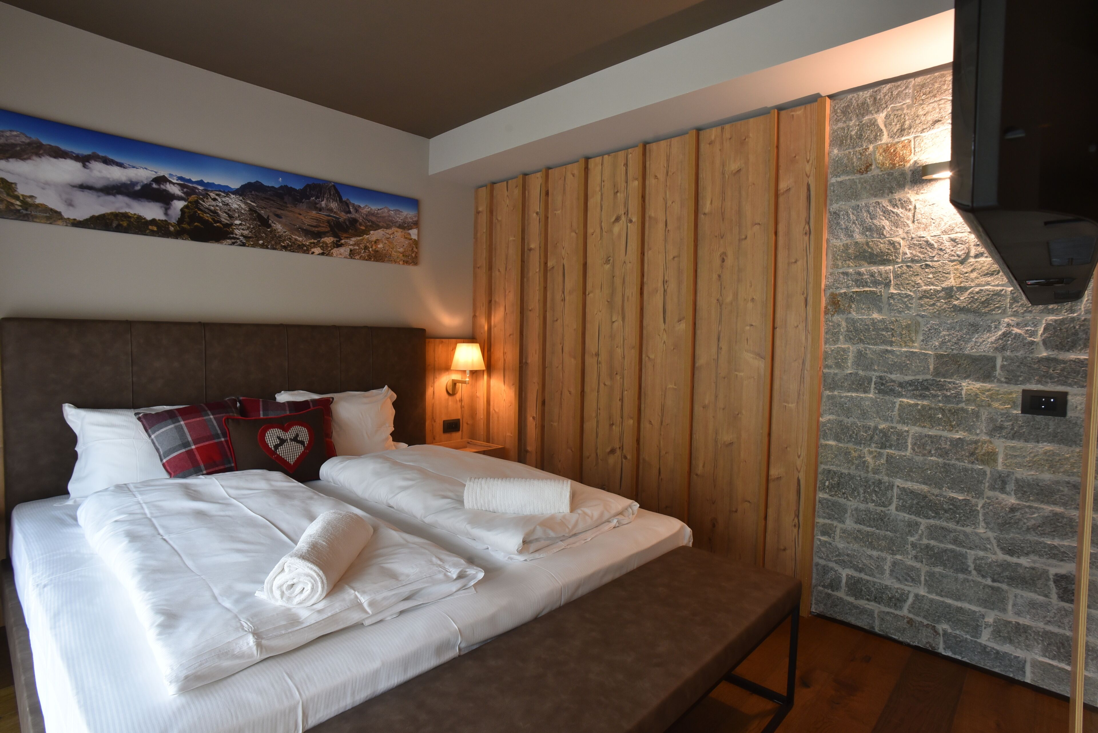 A hotel room with a bed, wooden panel wall, stone wall, and a mounted TV.