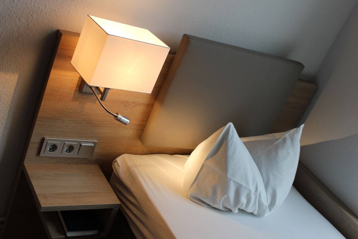 A neatly made bed with a pillow, a bedside lamp, and a wooden nightstand with a power outlet.